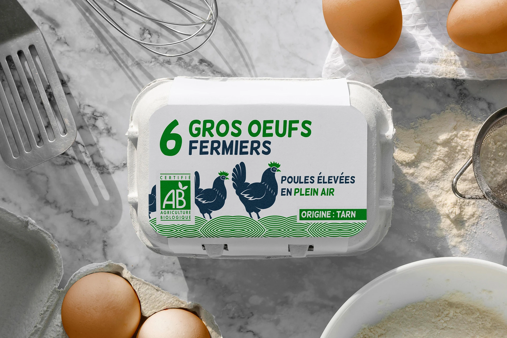 Étiquettes pour Boîtes à Œufs - Fromager à Toulouse Cette image montre une boîte de six gros œufs fermiers avec une étiquette indiquant 6 GROS OEUFS FERMIERS et POULES ÉLEVÉES EN PLEIN AIR. L'origine des œufs est mentionnée comme étant Tarn. L'étiquette affiche également le logo de la certification Agriculture Biologique (AB), signifiant que les œufs sont biologiques. La boîte est posée sur une surface en marbre entourée d'ustensiles de cuisson, d'œufs et de farine, suggérant un contexte de pâtisserie ou de cuisine. Cette image est pertinente pour la création et l'impression d'étiquettes pour boîtes à œufs pour un fromager basé à Toulouse, car elle montre un exemple clair de conception et de présentation d'étiquettes.